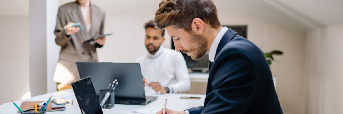 A businessman in a suit writing on a document at a desk while colleagues work on laptops in the background.