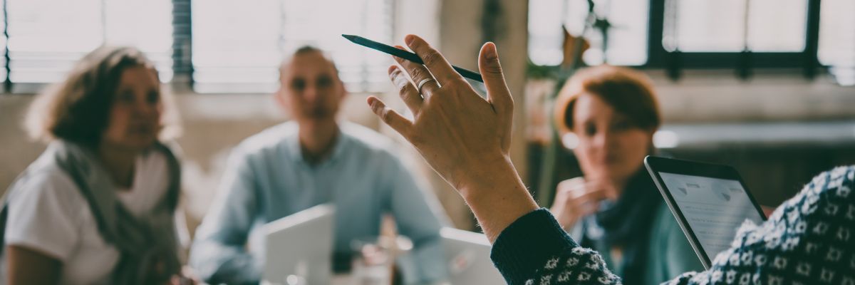 A close-up of a hand holding a pen during a meeting, with blurred colleagues and a tablet in the background.