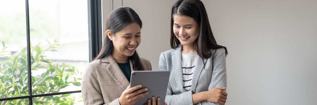 Two women in professional attire smiling while looking at content together on a tablet.