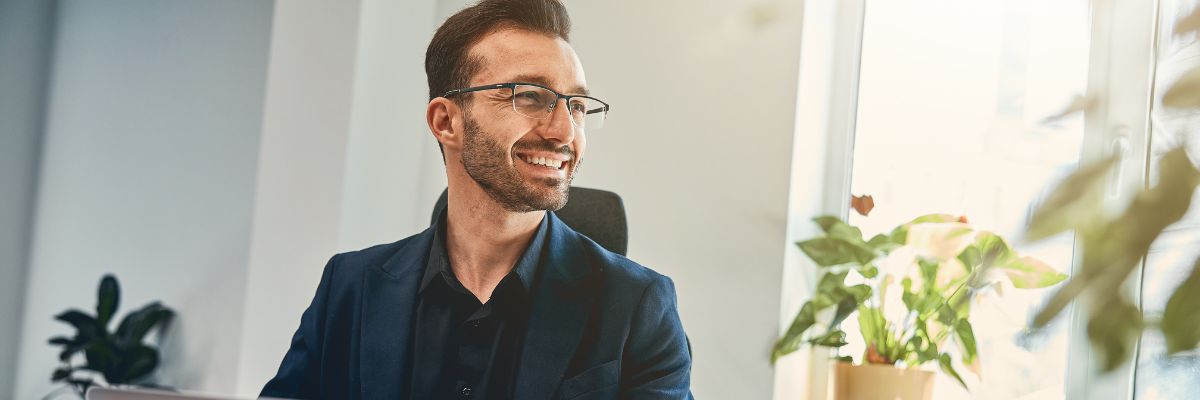 A smiling man in a suit looking out a bright office window.