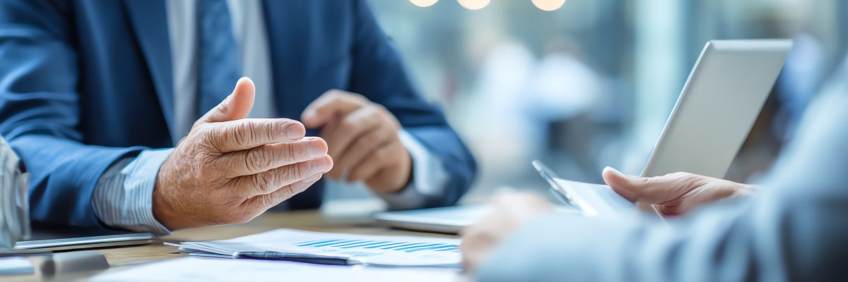 A close-up of two people in suits discussing financial charts at a desk, one gesturing while the other holds a tablet.